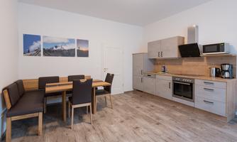 A modern kitchen with a dining area. The walls are light, and the furniture is made of wood and gray.