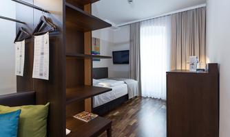 A modern room with a bed and a desk. There is wooden flooring, large windows, and a wardrobe.