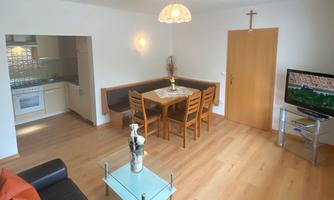 A bright living room with a dining table and chairs. In the background, an open kitchen and a TV can be seen.
