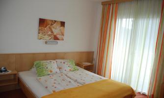 A cozy bedroom with a large bed and colorful curtains. A piece of artwork hangs on the wall, and plenty of daylight streams through the window.