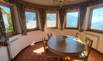A bright dining room with a round table and six chairs. Large windows offer a view of the surrounding mountains.