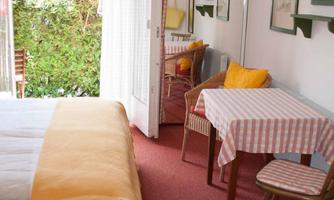 A bright room with a bed and a small table. In the background, chairs and a green view are visible.
