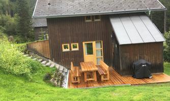 A cozy wooden house surrounded by green grass and trees. Outside, there is a wooden table set on the terrace.