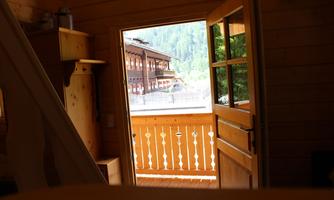 A cozy wooden door opens to a balcony overlooking a traditional Alpine house. In the background, green mountains can be seen.