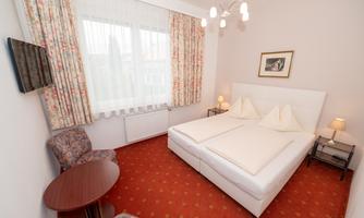 A cozy hotel room with a large double bed and a small table. The windows are decorated with curtains, and the room has a warm, inviting carpet.