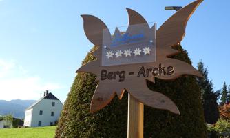 A wooden sign with the inscription "Mountain Ark" stands in front of a green bush. In the background, a white building and a blue sky are visible.