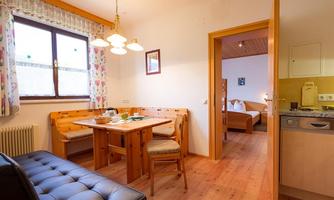 A cozy living space with a wooden dining table and a small kitchenette. In the background, a bedroom and a window with curtains are visible.