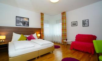 A modern bedroom with a double bed and colorful decorations. The room has large windows that let in a lot of light, and a cozy sitting area.