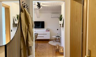 A modern hallway with wooden flooring and bright wall design. In the background, a living room with a TV and a cozy seating area is visible.