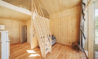 A modern, bright room with wooden paneling and a closed staircase. A sofa and a small table are available, as well as large windows that let in plenty of light.