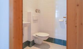 A modern bathroom with white tiles and a simple toilet. In the background, a sink and a small decorative element are visible.