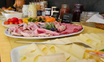 A buffet with various cold cuts, slices of cheese, and fresh fruit. In the background, jars of jam and other foods are visible.