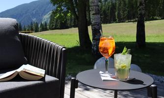 A cozy outdoor spot with two cocktails on a table. In the background, a green meadow and a picturesque mountain landscape stretch out.