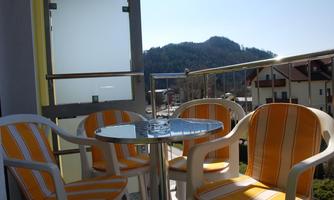 A balcony with two chairs and a small table. In the background, mountains and a clear blue sky are visible.