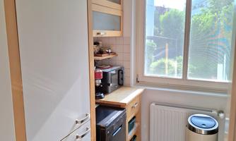 A modern kitchen with bright wood cabinets and large windows. The room features a refrigerator, microwave, and a trash can.