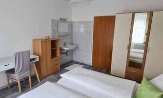 A simple bedroom with two beds and a desk. The walls are light, and there is a wardrobe as well as a bathroom.
