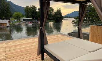 A cozy lounge platform on a wooden terrace overlooking a tranquil river. In the background, forests and mountains can be seen, while the sky displays soft colors.