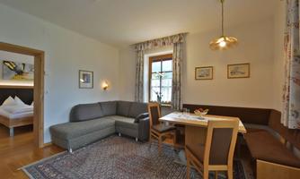 A cozy living room with a gray couch, a dining table, and wooden chairs. Large windows provide plenty of daylight.