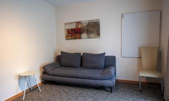 A cozy living room with a gray sofa and a small side table. On the wall hangs a modern artwork, and there is a bright chair in the corner.