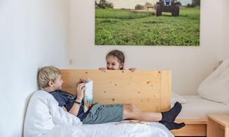 Two children are relaxing in a bright room. One is reading a book while the other is curiously looking over at the wall.