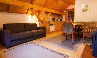 A cozy living room with wooden ceilings and a sofa. There is a dining table with chairs and a small kitchenette.