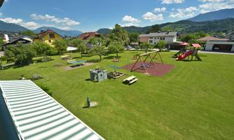 A spacious playground with swings and slides in a green meadow. In the background, hills and some houses can be seen.