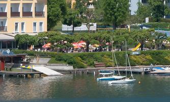 A tranquil lakeside landscape with a small boat on the water. In the background, houses and trees can be seen, as well as a bustling terrace.