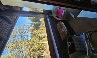 A cozy balcony with two chairs and a small table. In the background, there are tall trees and a clear blue sky.