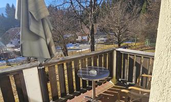 A sunny balcony with a table and two chairs. In the background, trees and a peaceful landscape can be seen.