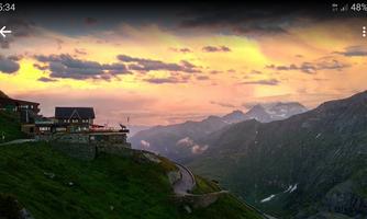 A stunning mountain landscape at sunset with a mountain cabin in the foreground. The clouds glow in soft colors over the peaks.