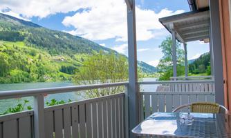 A cozy terrace with a table and chairs. In the background, you can see a beautiful view of a green lake and the surrounding mountains.