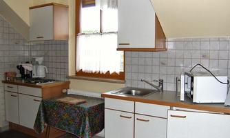 A small, bright kitchen with wooden cabinets and a windowsill. A colorful cloth is laid on the table, and various kitchen appliances are visible.