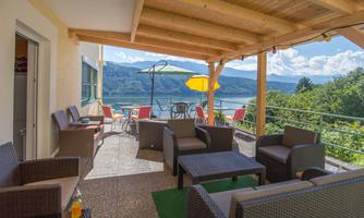 A cozy terrace with seating made of rattan and a beautiful view of the lake and the mountains. Umbrellas provide shade on a bright and sunny day.