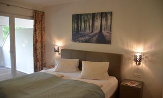 A cozy bedroom with a double bed and two pillows. There is a picture of a forest hanging on the wall, and there are windows overlooking the outside.