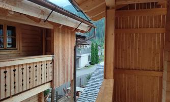 A wooden veranda overlooking a half-timbered house and a paved path. The surroundings are surrounded by nature and have a cozy atmosphere.