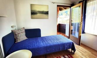 A simple room with a blue bed and a table. There is a window that leads to a balcony, and a picture on the wall.