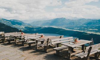 A terrace with wooden tables and benches offers a beautiful view of the mountains and the valley. The landscape is green and the weather is clear.