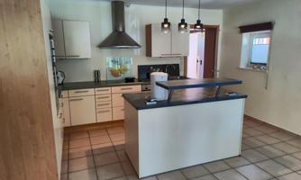 A modern kitchen with a central island and various cabinets. The walls are bright, and there are several lamps that illuminate the room well.