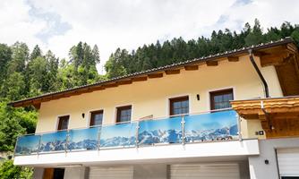 A modern house with a balcony and a glass railing that shows the mountains. The surrounding nature is green and mountainous.