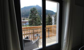 A view from a window onto a terrace with a table and a chair. In the background, trees and mountains are visible.