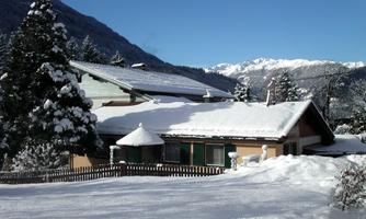 A snow-covered house in a winter landscape. The mountains in the background are clearly visible under a blue sky.