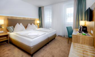 The image shows a modern hotel room with two beds, a cozy seating area, and a desk. Large windows allow plenty of natural light to come in.