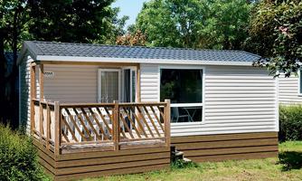 A modern holiday cottage with a wooden terrace and a large window front. It is located in a green environment with trees.