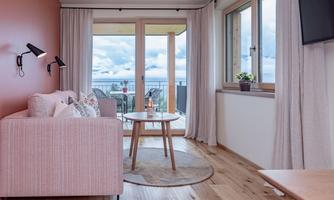 A modern living room with a pink sofa and a table. Large windows provide a view of the terrace and the sky.