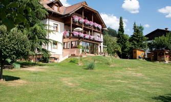 A pretty house with balconies decorated with flowers. The garden is green and surrounded by trees.