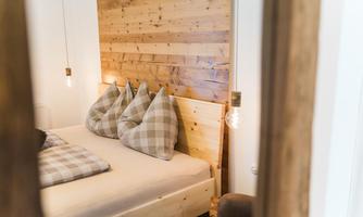 A cozy bedroom with a wooden bed and soft pillows. The wall is covered with wooden panels and warm light sources are hanging.