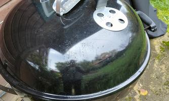 A round, black grill stands on the ground. In the background, wooden walls and a grill lid can be seen.