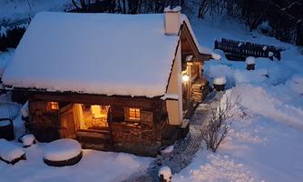 A charming wooden house in the snow, with warm light coming from the windows. The surroundings are covered with a thick layer of snow, creating a tranquil winter landscape.