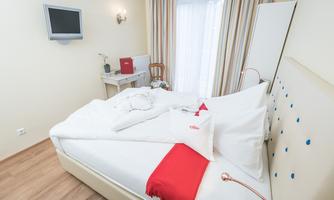 A cozy room with a double bed and a light wooden floor. There is a desk, a television, and plenty of daylight.