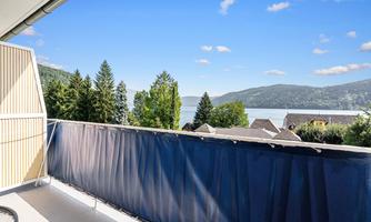 A sunny view from a balcony overlooking a clear lake surrounded by trees. The sky is blue and there are a few clouds visible.